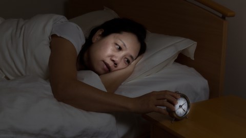 Woman with ADHD is sleepless touching alarm clock while eyes open.