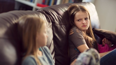 Two young girls sit on couch looking annoyed at each other.
