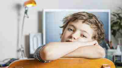 Boy with ADHD folds arms over back of chair in front of work desk looking bored